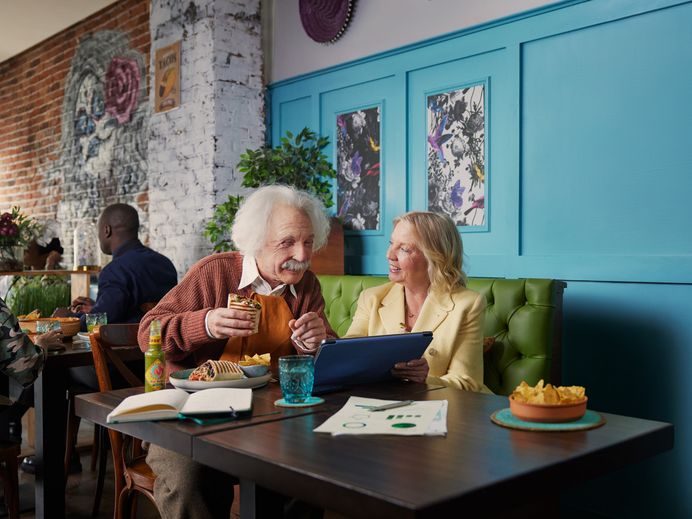 Elderly man styled as Albert Einstein sits with Deborah Meaden in a Mexican restaurant looking at a computer tablet with business paperwork nearby.